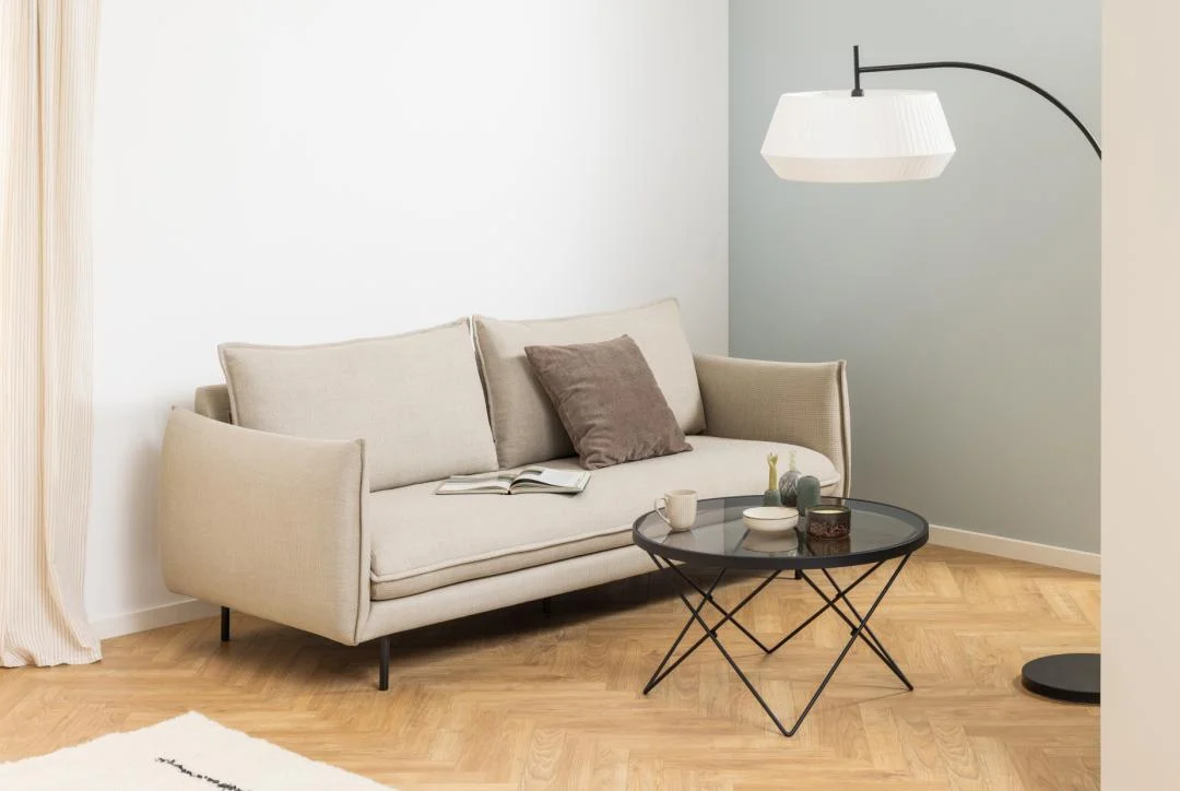 Een moderne woonkamer met een beige bank, bruin kussen, open boek en de Salontafel Zwart, Grijs 80 CM Rond Staal salontafel met planten en kaarsen, plus een vloerlamp met een witte kap op een houten visgraatvloer.