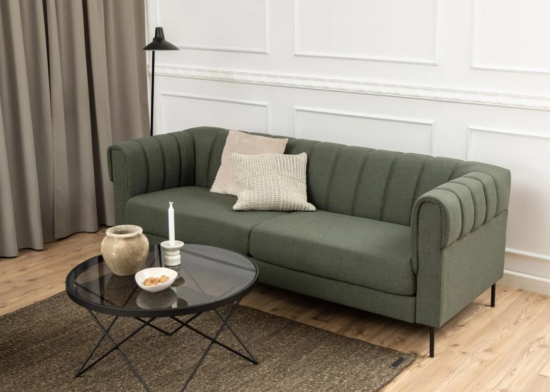 Een moderne woonkamer met een groen gestoffeerde bank, twee kussens, een Salontafel Zwart, Grijs 80 CM Rond Staal bekleed met decor, een bruin vloerkleed, staande lamp en beige gordijnen tegen een wit gelambriseerde muur en houten vloer.