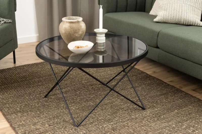 De Salontafel Zwart, Grijs 80 CM Rond Staal staat op een bruin geweven vloerkleed, gestileerd met een beige vaas, een witte kandelaar en kaars en een witte schaal, geplaatst tussen groene banken.