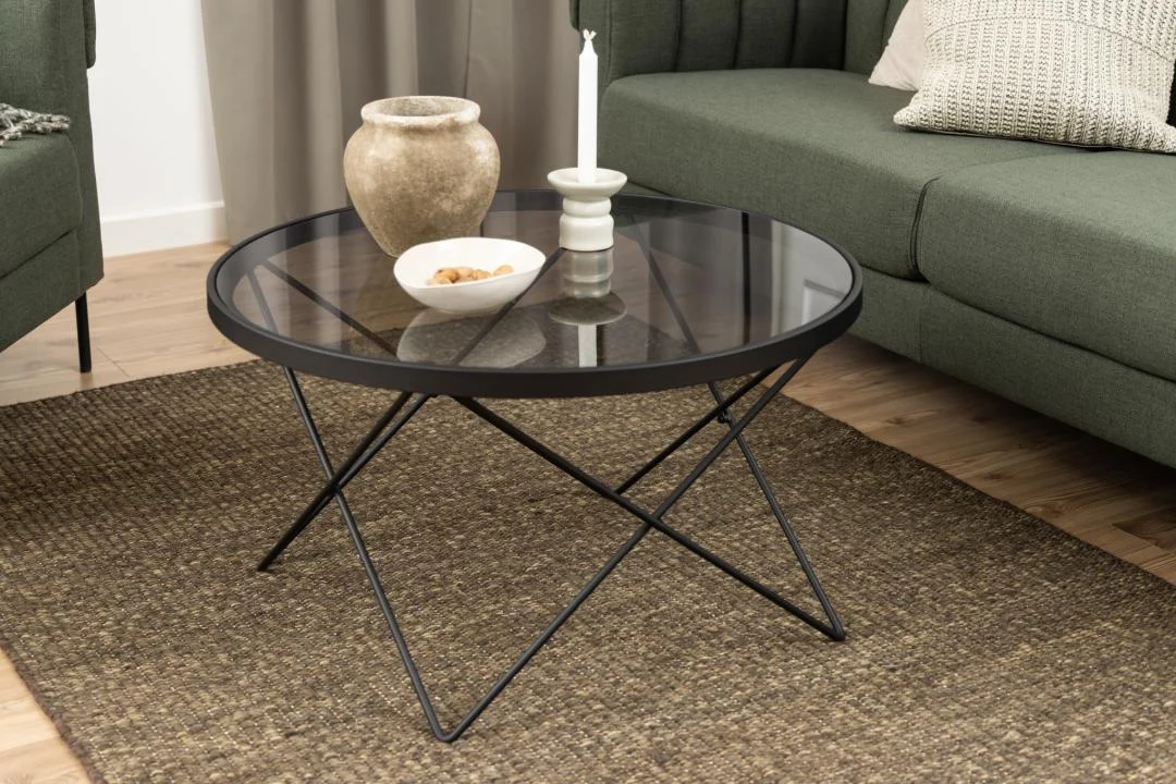 De Salontafel Zwart, Grijs 80 CM Rond Staal staat op een bruin geweven vloerkleed, gestileerd met een beige vaas, een witte kandelaar en kaars en een witte schaal, geplaatst tussen groene banken.