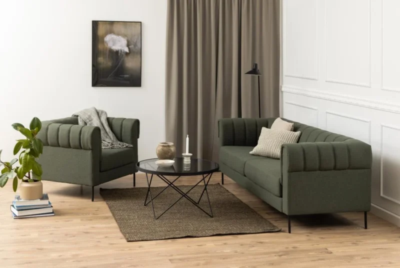 Een moderne woonkamer heeft een groene bank en fauteuil, een beige vloerkleed, een plant in pot, een staande lamp, neutrale gordijnen en witte wanden met panelen. De minimalistische inrichting wordt compleet gemaakt door de salontafel Zwart, Grijs 80 CM Rond Staal.