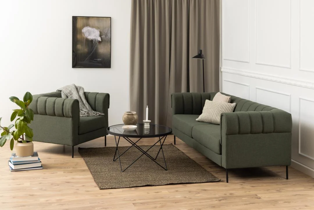 Een moderne woonkamer heeft een groene bank en fauteuil, een beige vloerkleed, een plant in pot, een staande lamp, neutrale gordijnen en witte wanden met panelen. De minimalistische inrichting wordt compleet gemaakt door de salontafel Zwart, Grijs 80 CM Rond Staal.