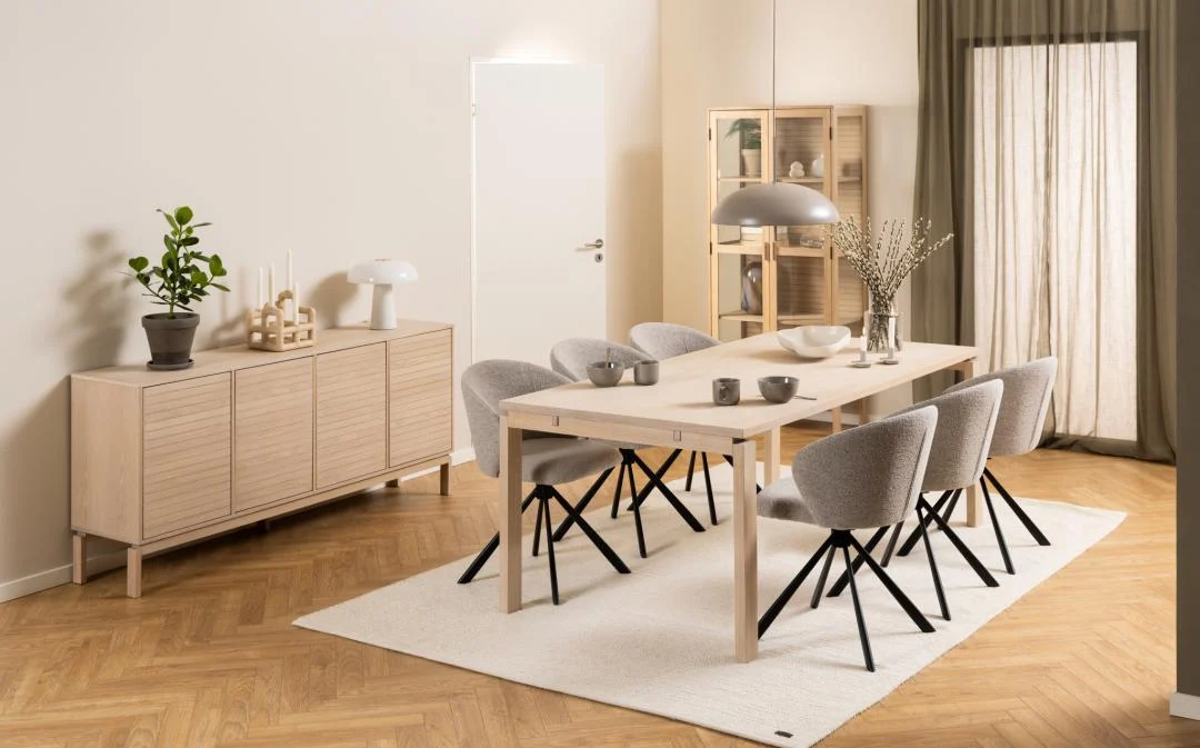 Minimalistische eetkamer met de Eettafel Nieuwe Sfeer 100 x 75 CM, een rechthoekige eiken tafel, zes grijze stoelen met kussens, een wit vloerkleed, een dressoir met planten en decor, een glazen kast, een houten vloer en zacht natuurlijk licht uit een raam met gordijnen.
