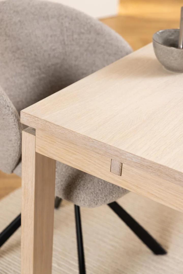 Close-up van de Eettafel Nieuwe Sfeer (100 x 75 CM), een rechthoekige eiken eettafel, met vierkante scharnierdetails. Ernaast staat een grijze stoel met kussens en zwarte poten; een kom en lepel rusten gedeeltelijk op de tafel.