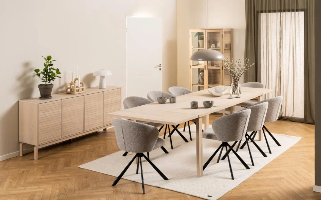 In een moderne eetkamer staat de Eettafel Nieuwe Sfeer (rechthoek eikenhout, 100x75 cm) met acht grijs gestoffeerde stoelen op een wit vloerkleed, een gedecoreerd dressoir, hanglamp boven en een glazen kast bij grote ramen met transparante gordijnen.