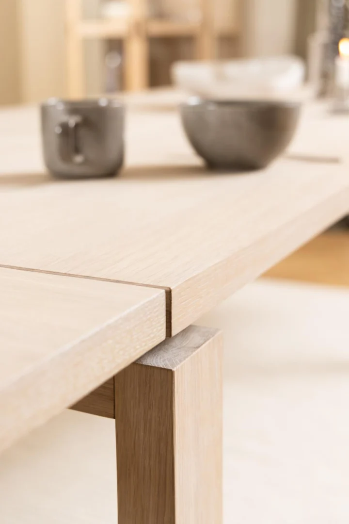 Close-up van de Eettafel Nieuwe Sfeer (100 x 75 CM), een rechthoekige eikenhouten eettafel met precies in elkaar gezette hoeken. Een grijze kop en kom rusten op het tafelblad, tegen een zacht vervaagde, gezellige achtergrond binnenshuis.