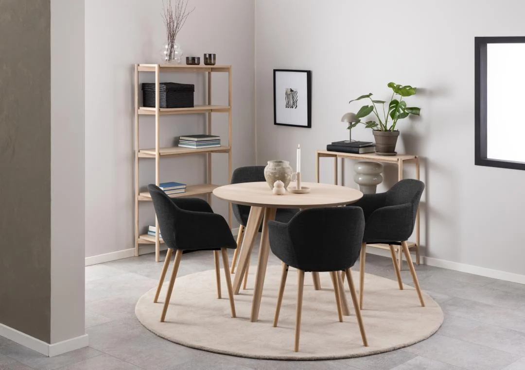 In een moderne eetkamer staat de Eettafel Townsville 100 CM Rond Eikenhout met vier zwart gestoffeerde stoelen op een crèmekleurig vloerkleed. Vlakbij staan twee lichthouten planken met boeken, planten en decor tegen lichtgrijze muren.