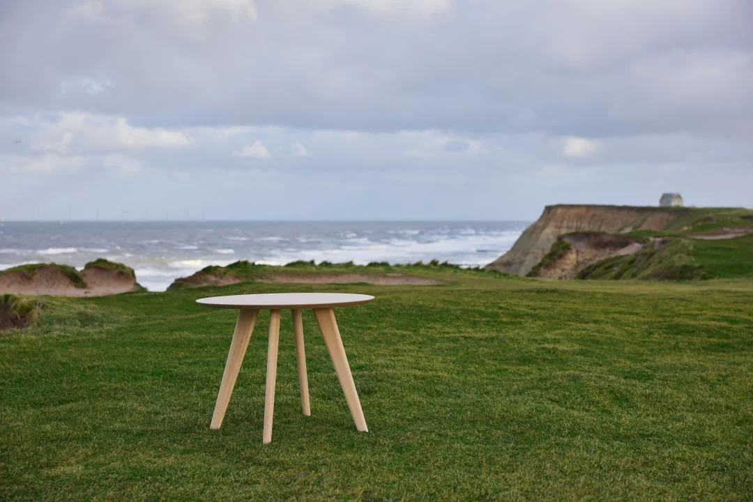 De Eettafel Townsville 100 CM Rond Eikenhout staat op groen gras bij een kustklif, met de oceaan en een bewolkte lucht op de achtergrond.