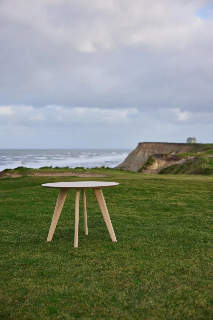 De Eettafel Townsville 100 CM Rond Eikenhout, een compacte ronde eiken tafel met lichte poten, staat op een grasveld bij kliffen met uitzicht op de oceaan onder een bewolkte hemel.
