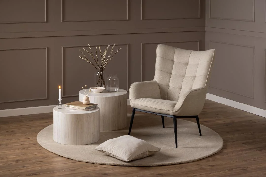Een beige fauteuil staat op een Salontafel Beige, Roest 40 CM Rond - een elegante ronde beige plastic tafel - naast decorstukken in een zacht verlichte, neutraal gekleurde kamer. Een beige kussen rust op de vloer.