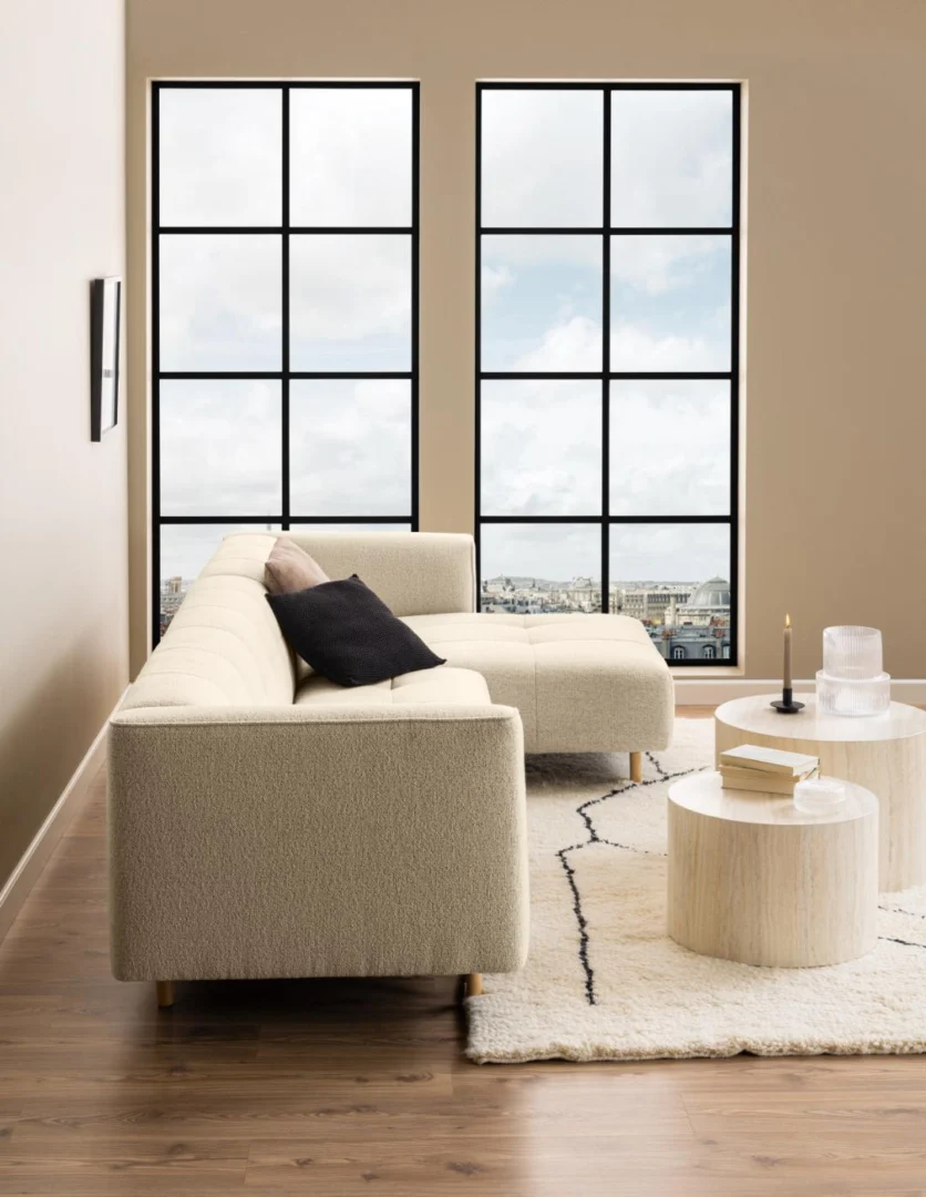 Een moderne woonkamer is voorzien van een beige sectionele bank, twee salontafels Beige, Roest 40 CM Rond in kunststof, zwarte en beige kussens, een licht vloerkleed en grote rasterramen met uitzicht op een bewolkt stadslandschap.