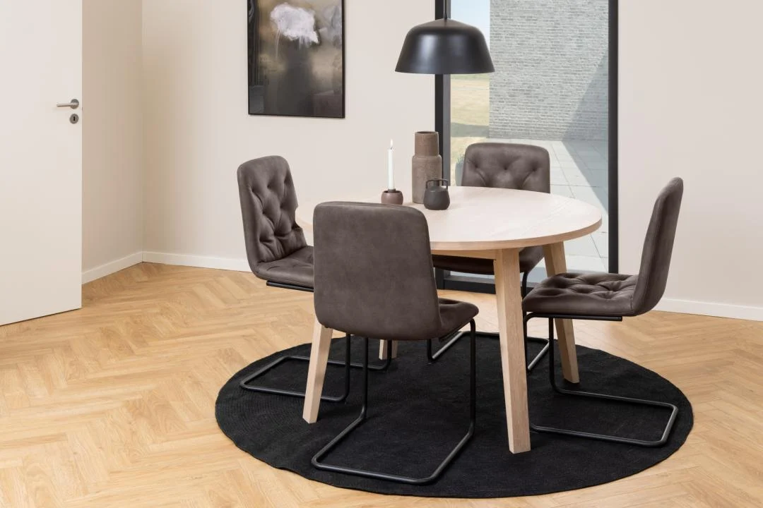 Een moderne eethoek met de Eettafel 120 CM Rond Eikenhout, vier bruine stoelen met kussens, een zwarte hanglamp en tafeldecor, allemaal gerangschikt op een zwart cirkelvormig tapijt op een lichthouten vloer.