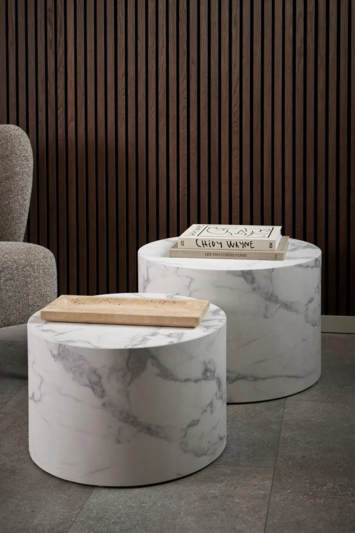 Twee Salontafels Wit 40 CM Rond Kunststof staan op een grijze tegelvloer. Op de ene staat een rechthoekig houten dienblad, op de andere twee gestapelde boeken. Op de achtergrond staan een gestructureerde fauteuil en een donkerhouten lattenwand.