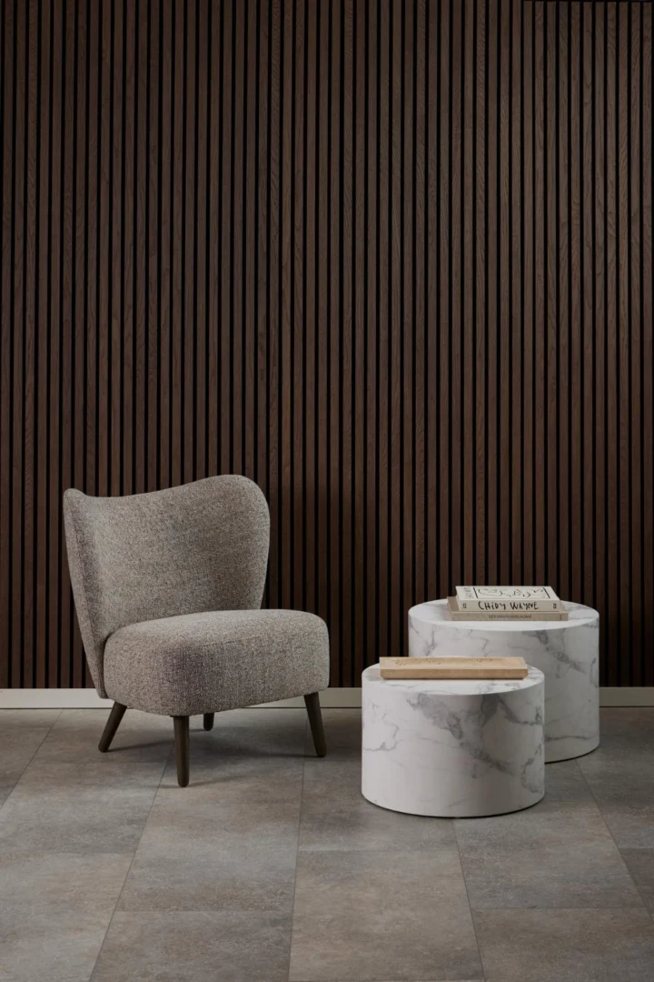 Een grijs gestoffeerde fauteuil staat naast twee Salontafel Wit 40 CM Rond Kunststof tafels voor een verticale donkerhouten lattenwand, met een krant en een boek op de tafels.