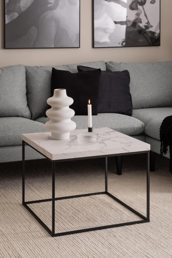Een moderne woonkamer met een lichtgrijze sectionele bank, de Salontafel Zwart, Wit 60 x 60 CM Vierkant in marmerlook met een zwart frame en wit bovenblad (kunststof), een sculpturale vaas, een kaars en abstracte muurkunst op de achtergrond.
