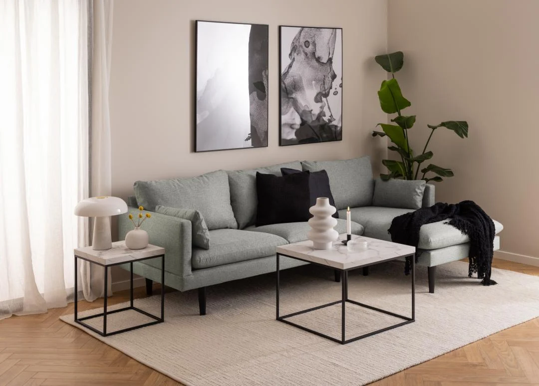 Een moderne woonkamer met een lichtgrijs bankstel, zwart-witte abstracte kunst, een grote groene plant, een neutrale inrichting, een licht vloerkleed op een houten vloer en een Salontafel Zwart, Wit 60 x 60 CM Vierkant (kunststof) als salontafel.