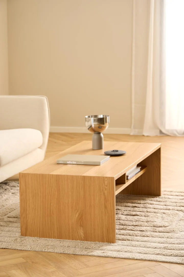 In een minimalistische woonkamer staat de Salontafel Roest 60 x 40 CM Rechthoek in eikenfineer op een vloerkleed met structuur, afgewerkt met een metalen schaal, kleine vaas, boek en afstandsbediening. Een crèmekleurige bank en transparante gordijnen zorgen voor een zachte achtergrond.