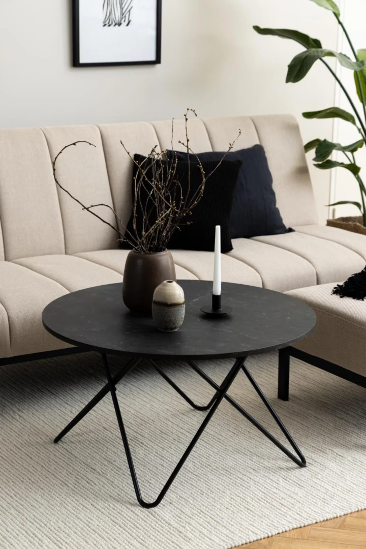Een moderne woonkamer met een beige bank, zwarte en marineblauwe kussens en een compacte Salontafel Zwart 80 CM Rond Kunststof met een bruine vaas met takjes, een kleine keramische pot en een zwart-witte kandelaar; op de achtergrond staat een plant.