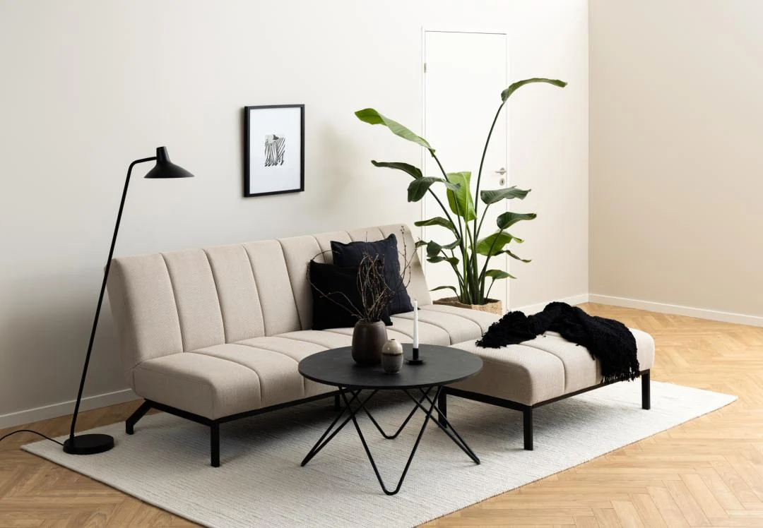 Een moderne woonkamer met een beige bank, zwarte staande lamp, Salontafel Zwart 80 CM Rond Kunststof als compacte ronde salontafel, groene plant in pot, ingelijste muurkunst, licht vloerkleed op houten vloer en zwarte plaids en kussens voor het contrast.