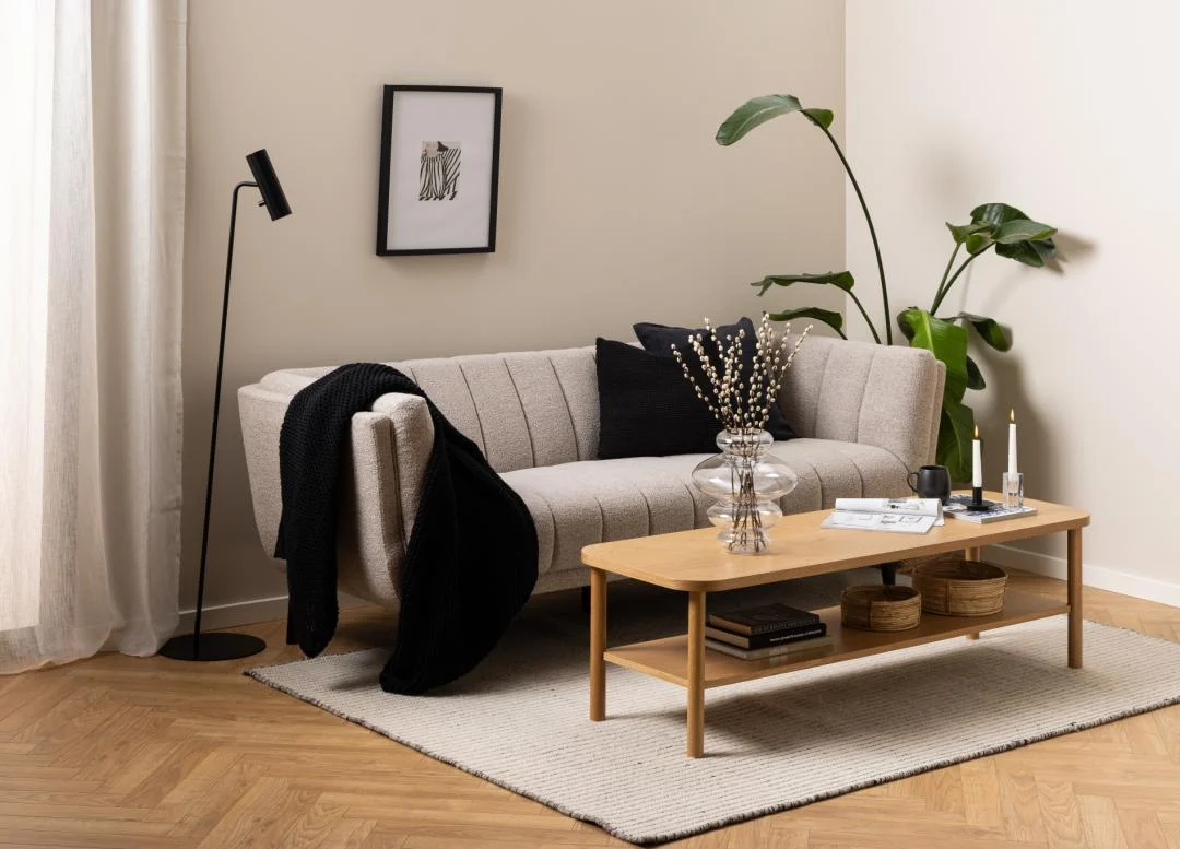 Een moderne woonkamer met een beige bank, zwarte en beige kussens, een zwarte staande lamp, ingelijste muurkunst, een grote groene plant en de Salontafel Roest 134 x 34 CM Rond in eikenfineer op een licht vloerkleed over een houten vloer.