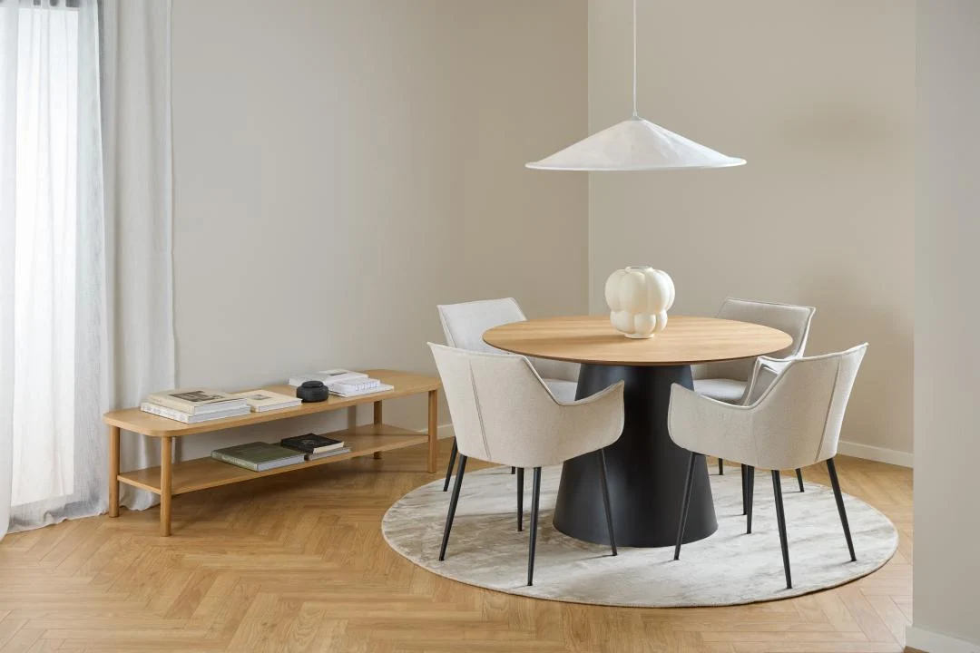 In een moderne eethoek staat de Salontafel Roest (134 x 34 cm, rond) in eikenfineer, omringd door vier beige gestoffeerde stoelen, een witte hanglamp, een witte vaas op tafel en een licht rond vloerkleed op een vloer van visgraathout.