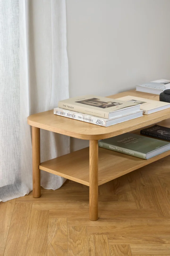 De Salontafel Roest, een ronde salontafel in eikenfineer van 134 x 34 cm, biedt plaats aan netjes gestapelde boeken en tijdschriften. Hij staat op een houten vloer bij een wit gordijn en een lichtgekleurde muur.