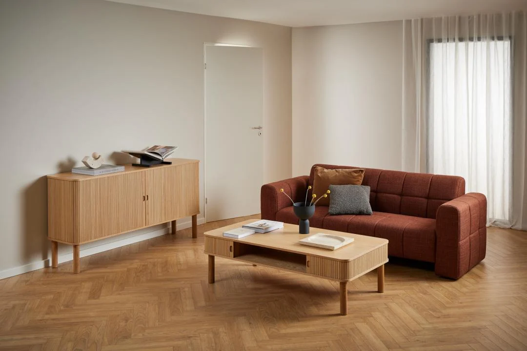 Een moderne woonkamer met lichthouten vloeren, een roestkleurige bank en kussens, de Salontafel 70 x 40 CM Rond Eikenfineer met daarop boeken en een vaas, plus een dressoir met decor, allemaal tegen crèmekleurige muren en transparante gordijnen.
