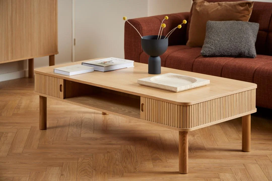 De Salontafel 70 x 40 CM Rond Eikenfineer met twee boeken, een dienblad en een zwarte vaas met gele bolbloemen staat op een houten vloer voor een roodbruine sofa met grijze en bruine kussens.