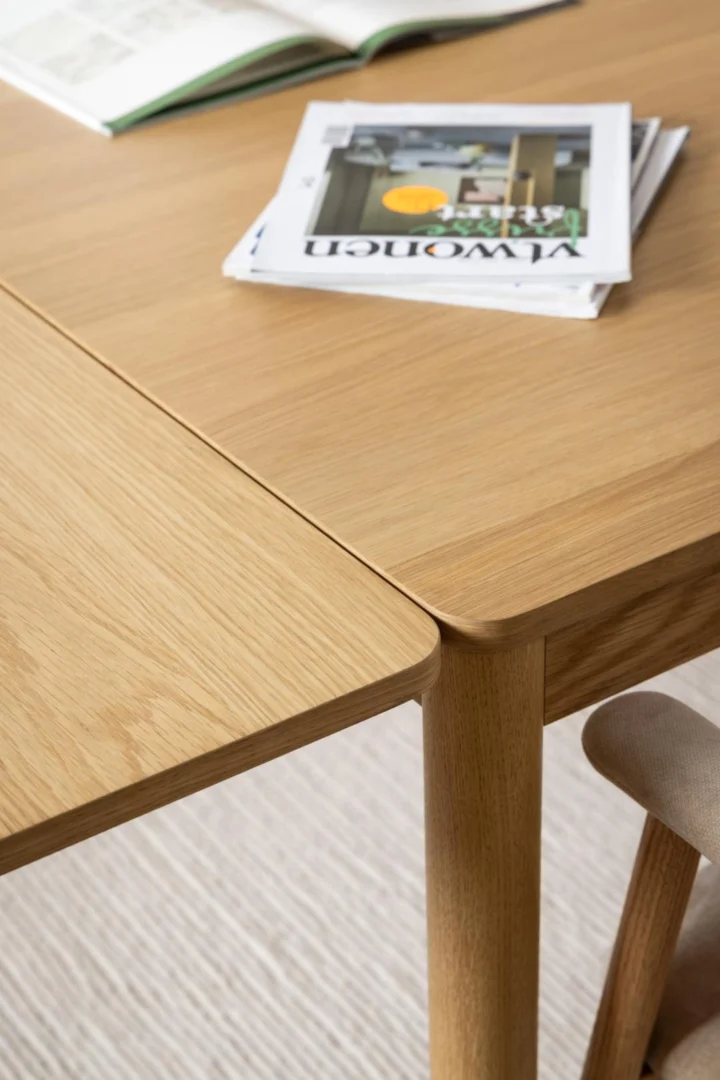 Een close-up van de Eettafel Nieuwe Sfeer 100 x 75 CM Rond, gemaakt van eikenhout, met een open boek en tijdschrift erop, naast een bijpassende houten stoel op een tapijt met een lichte structuur.