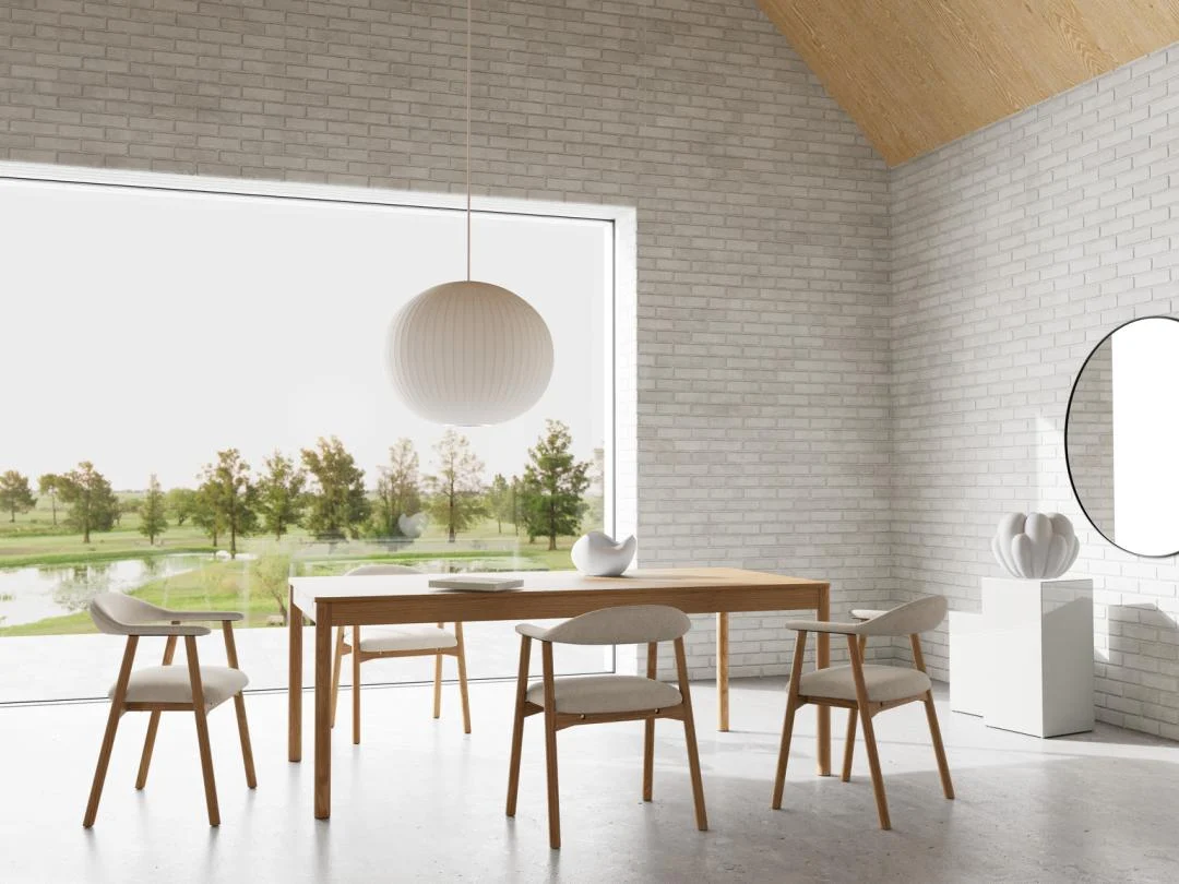 Minimalistische eetkamer met de Eettafel Nieuwe Sfeer 100 x 75 CM Rond in eikenhout, vier stoelen, een grote ronde hanglamp en decoratieve vazen. Een groot raam kijkt uit over bomen en een vijver. De ruimte heeft witte bakstenen muren en een gewelfd houten plafond.