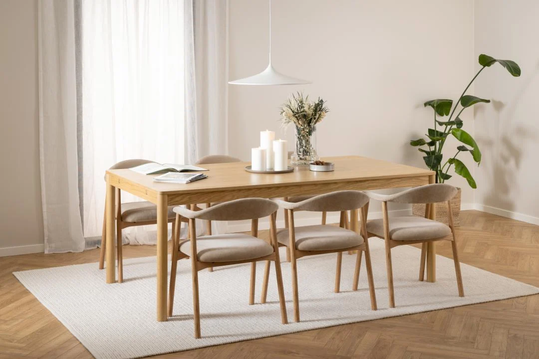 In een moderne eetkamer staat de Eettafel Nieuwe Sfeer 100 x 75 CM Rond in eikenhout, omringd door zes beige stoelen, met kaarsen, een vaas met droogbloemen, een open boek, een hanglamp en een groene plant op een wit vloerkleed op een houten vloer.