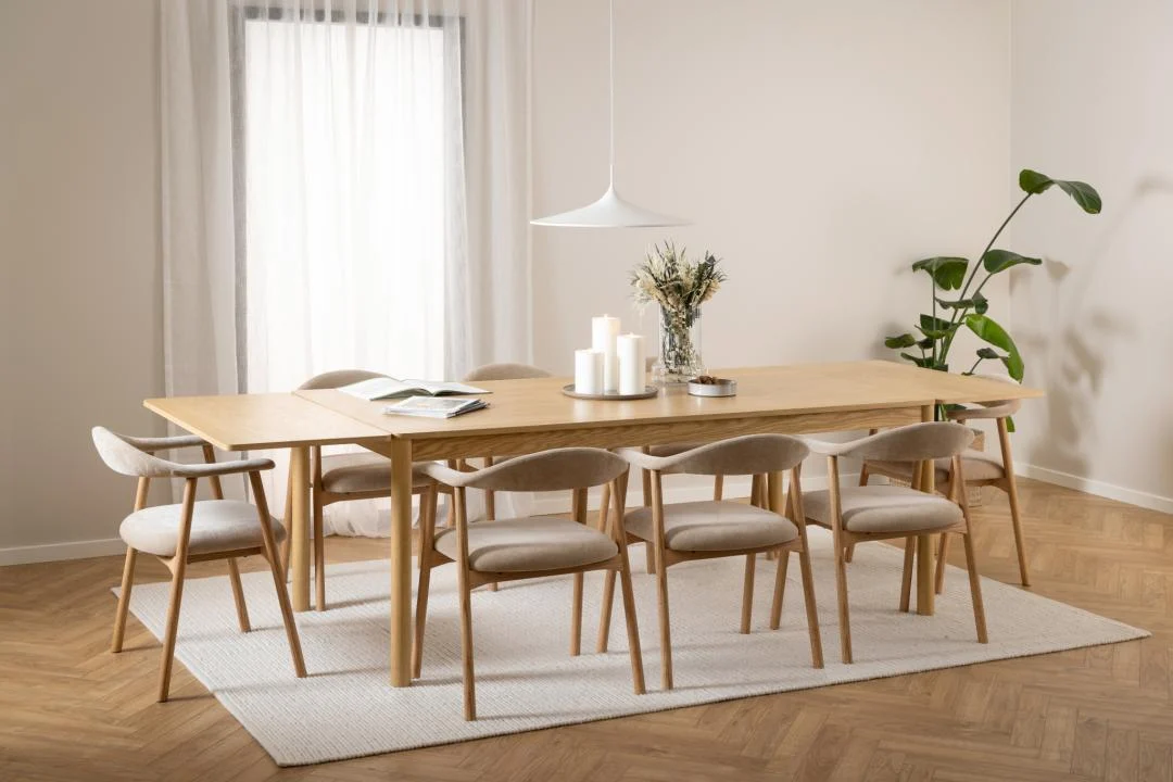 Een moderne eetkamer met de Eettafel Nieuwe Sfeer 100 x 75 CM Rond in eikenhout, acht stoelen met kussens, witte kaarsen, een vaas met droogbloemen, een hanglamp, een plant in pot en een groot raam met transparante gordijnen op een neutraal vloerkleed.