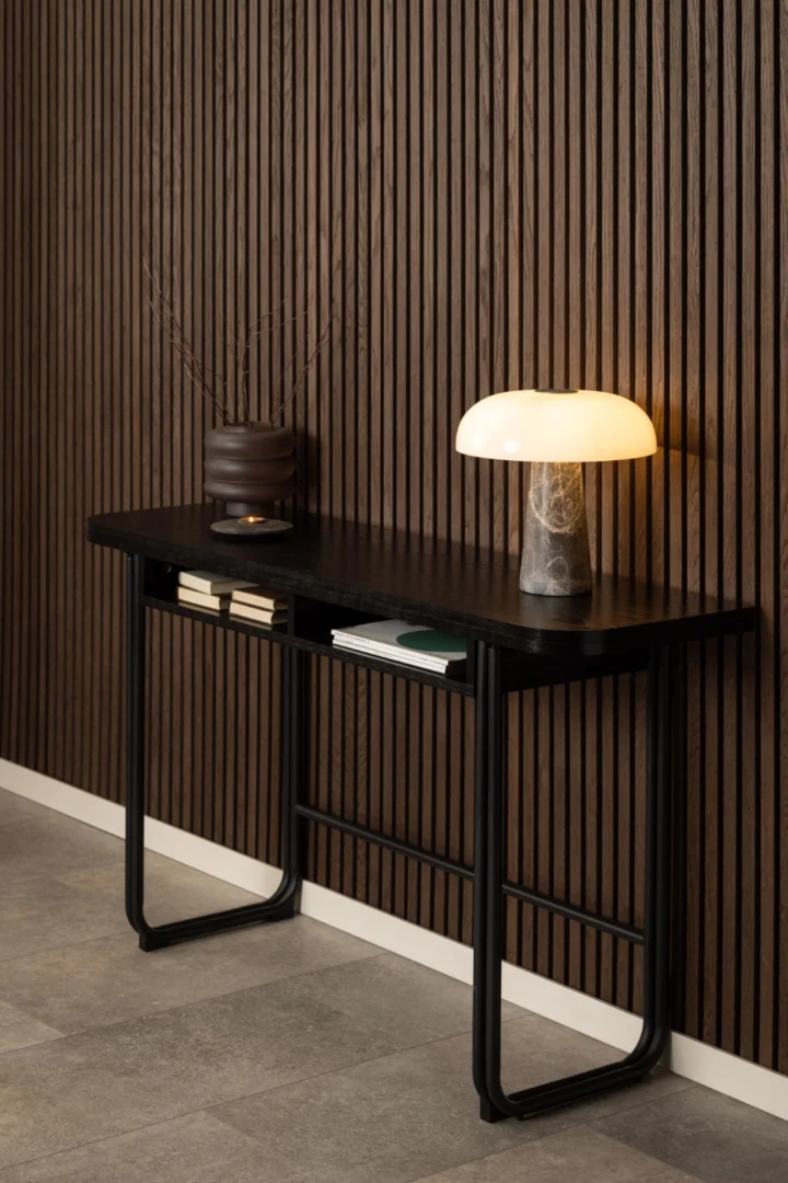 De Eettafel Nieuwe Sfeer (80 x 77 cm) van zwart MDF en staal toont boeken, een donkere vaas met takjes en een lamp van marmer met een gloeiende lampenkap, voor een verticale wand van houten latten en een grijze tegelvloer.