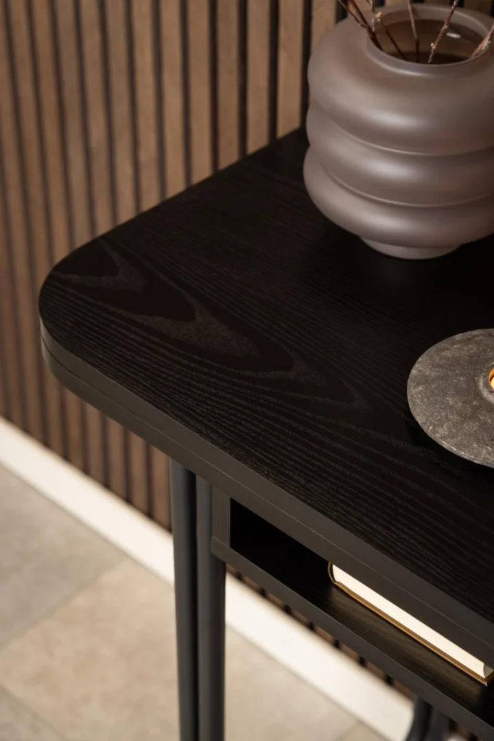 Een close-up van de Eettafel Nieuwe Sfeer (80 x 77 CM) toont het zwarte Rechthoek MDF blad en stalen frame, met afgeronde hoeken, versierd met een keramieken vaas en stenen onderzetter. Een houten lattenwand en tegelvloer verschijnen op de achtergrond.