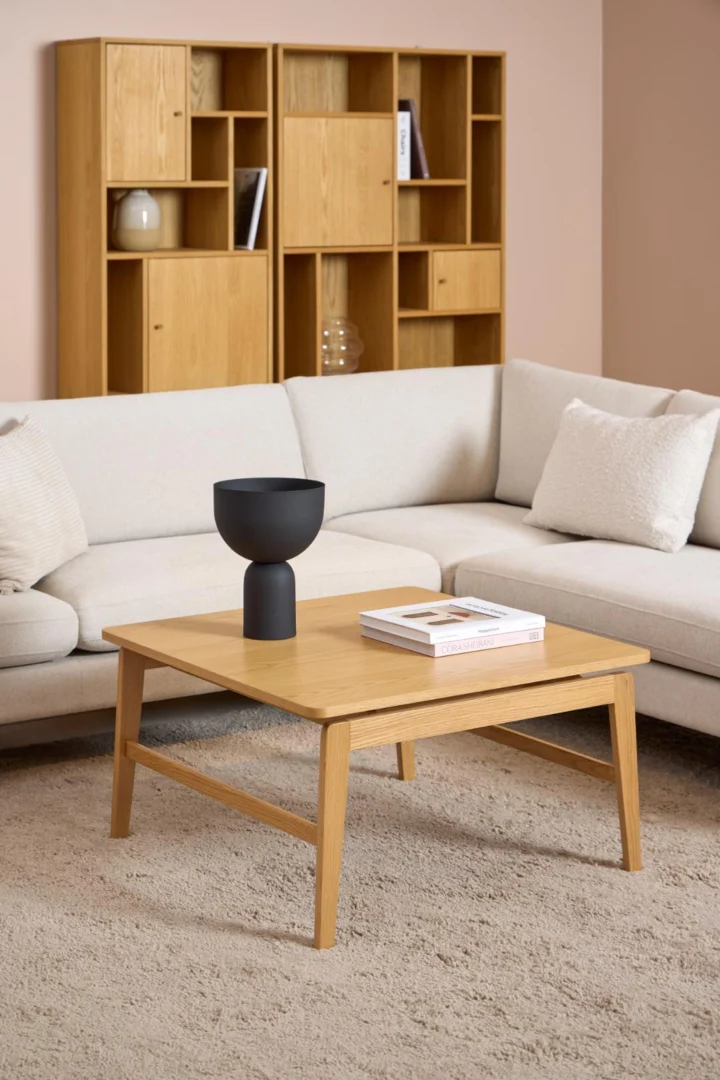 Een moderne woonkamer met een lichtbeige sofa, een 80 x 80 cm rechthoekige Salontafel van eikenfineer met daarin een zwarte vaas en twee boeken, een licht tapijt en houten rekken tegen een lichtroze muur.