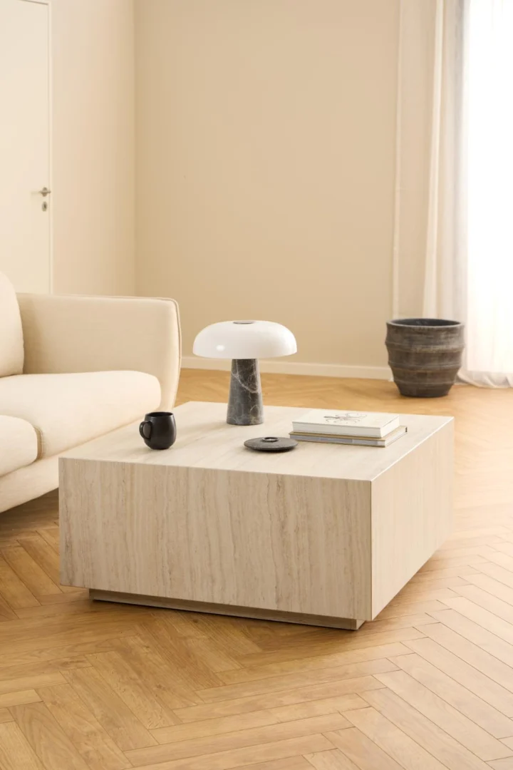 Een minimalistische woonkamer heeft een beige bank en de Salontafel Beige 90 x 42 CM Vierkant, een vierkante kunststof salontafel met een zwarte mok, boeken en een moderne witte lamp; natuurlijk licht stroomt binnen door witte gordijnen.