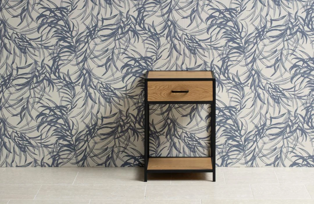Het Nachtkastje Zwart Eikenhout Industrieel (63 x 42 x 35 cm), met een zwart metalen frame en een enkele lade, staat tegen een witte muur met een blauw bladmotief en een lichtgekleurde tegelvloer.