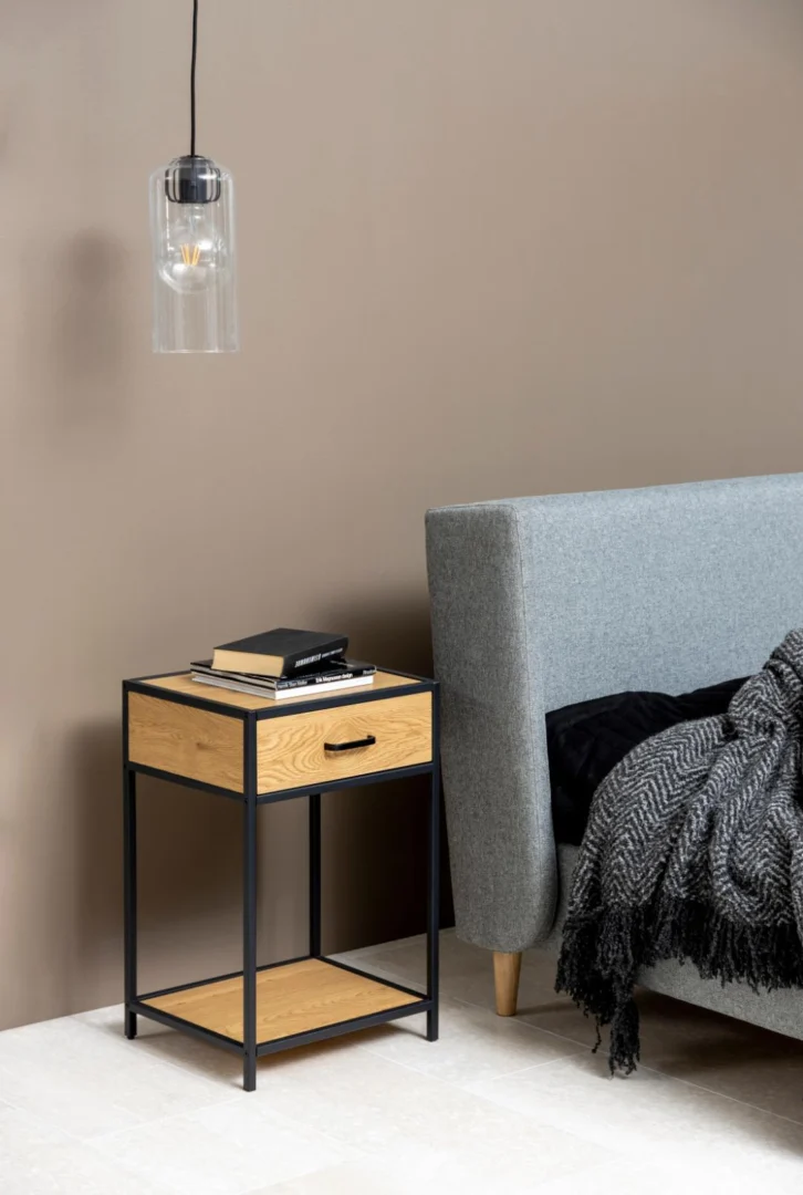 Een moderne slaapkamer heeft een grijs gestoffeerd bed, het Nachtkastje Zwart Eikenhout Industrieel (63x42x35 cm) met boeken erop, een hangende glazen hanglamp en een donker getextureerd deken gedrapeerd over het bed.