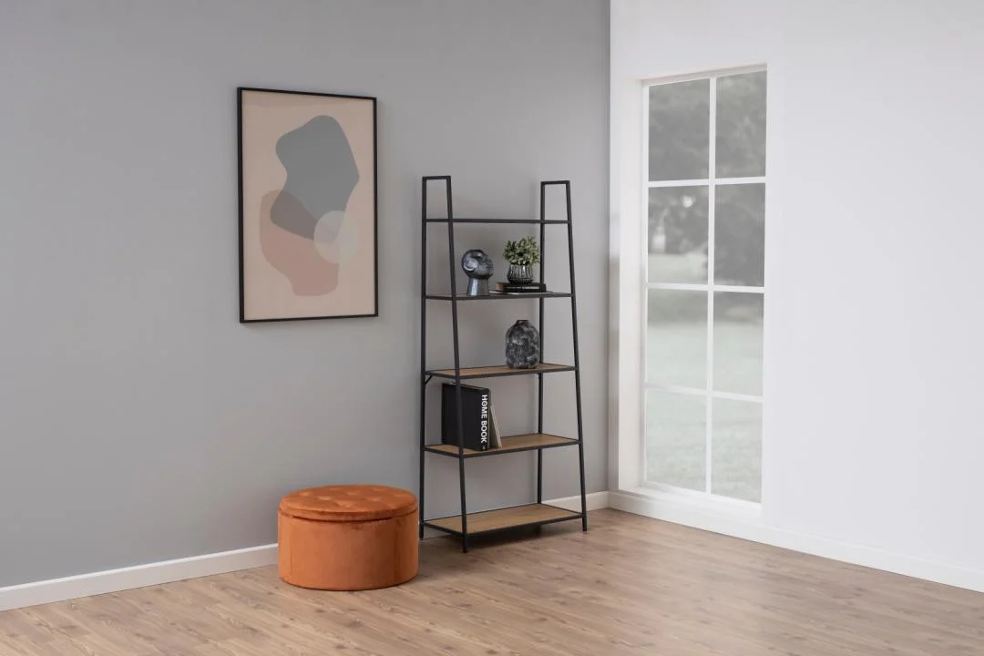 Een minimalistische kamer met lichte houten vloeren toont de Seaford boekenkast in zwart en hout, versierd met accenten, naast een abstract ingelijst kunstwerk op een grijze muur, een oranje voetenbankje en een groot raam.