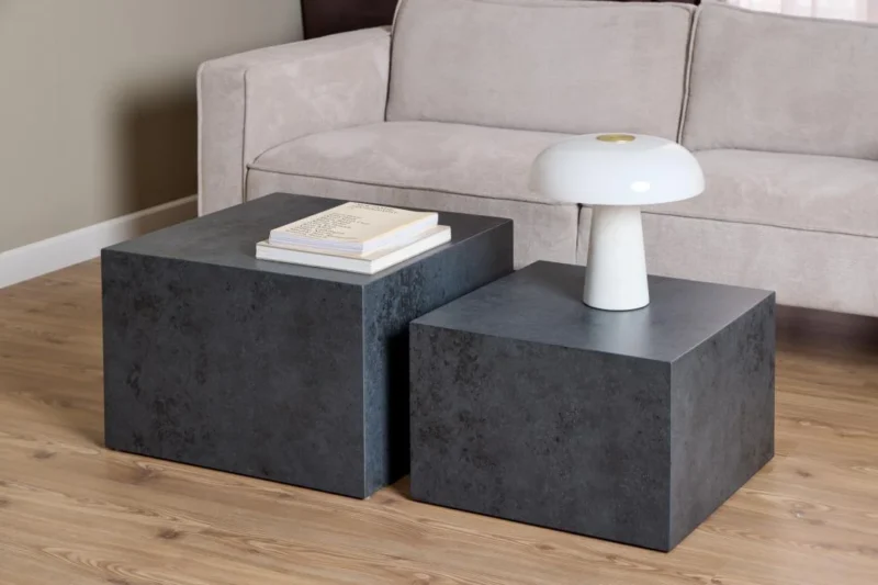 Twee Salontafel Zwart 58 x 40 CM Vierkant salontafels uit de Kunststof Nesting Set, de ene met een stapel boeken en de andere met een witte moderne lamp, staan voor een beige bank op een houten vloer.