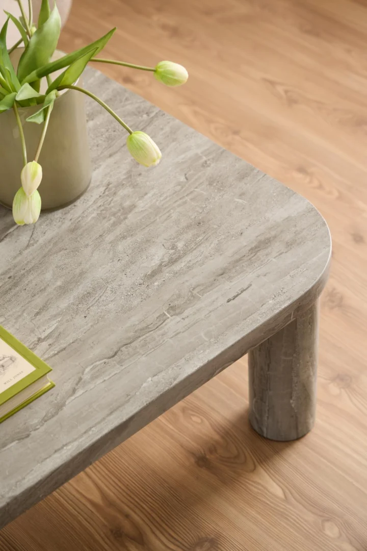 Een close-up van de Salontafel 75 x 36 CM Rond MDF, een moderne salontafel in grijze steenlook met afgeronde hoeken en poten op een lichte houten vloer, met een groene vaas met witte tulpen en een groen boek.