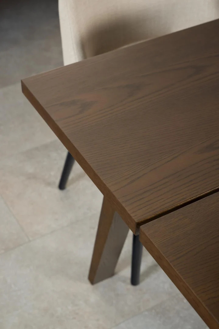 Een close-up van de Eettafel Nieuwe Sfeer 200 x 95 CM, een rechthoekige eiken eettafel met schuine poten, gekoppeld aan een beige gestoffeerde stoel op een lichtgekleurde tegelvloer.