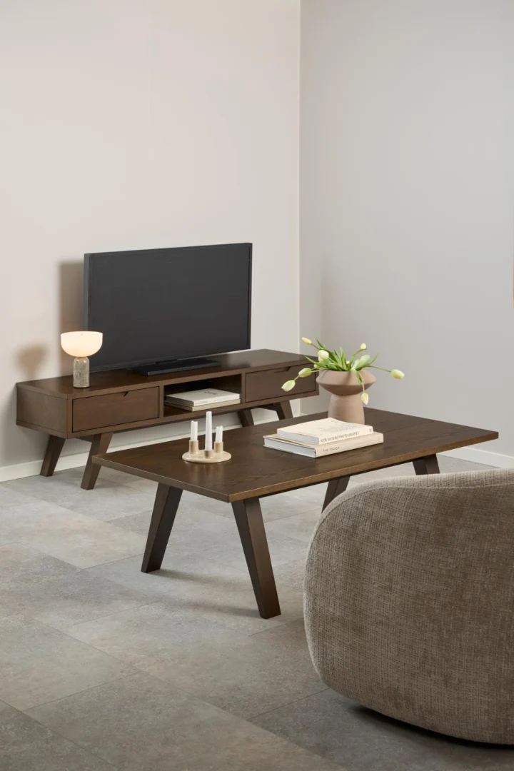 In een moderne woonkamer staan de Salontafel Grijs 70 x 45 CM Rechthoek in eikenfineer, een tv op een bijpassende houten standaard, een beige fauteuil, tulpen in een vaas, kaarsen, boeken en een lamp op een lichte tegelvloer tegen witte muren.