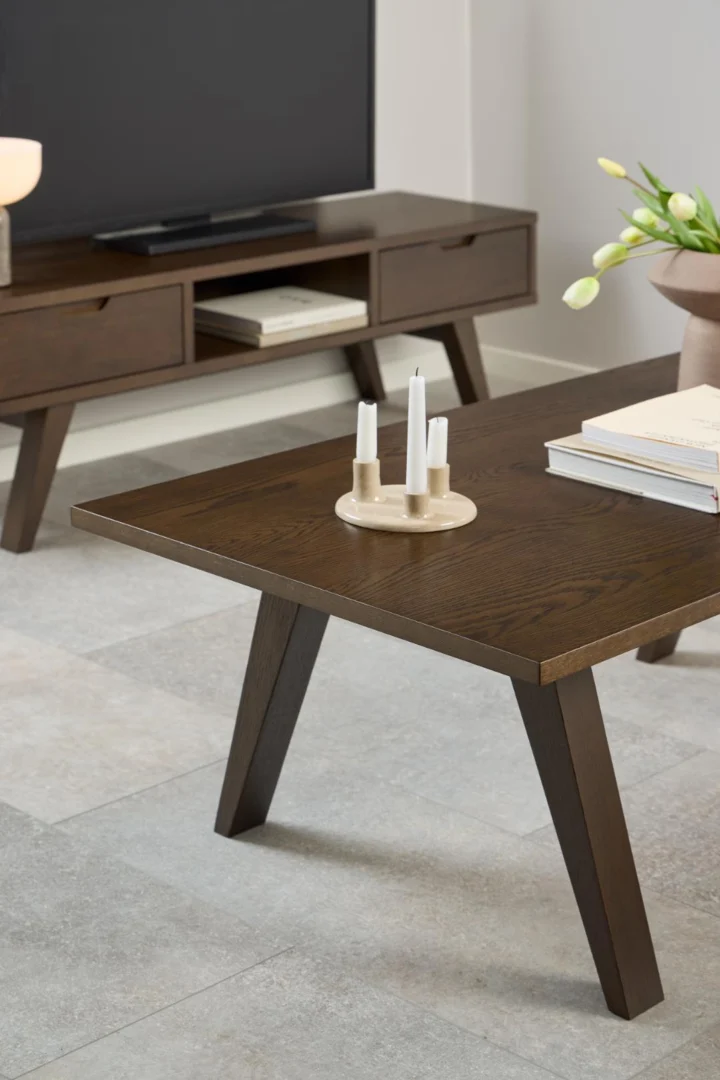 Een moderne woonkamer met een Salontafel Grijs 70 x 45 CM Rechthoek in eikenfineer, gecombineerd met een bijpassende TV-standaard. Op de tafel staat een kandelaar met drie witte kaarsen, boeken en er staat een vaas met witte tulpen op de achtergrond.