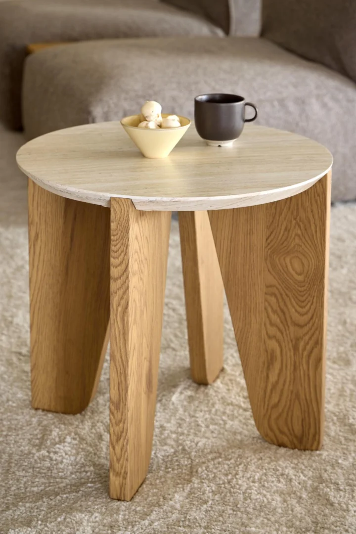 De Bijzettafel Beige 50 CM Rond Kunststof, een kleine ronde bijzettafel met drie schuine poten, bevat een bruine mok en een gele schaal met witte hapjes, geplaatst op een licht tapijt in de woonkamer.