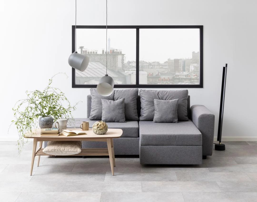 Een moderne woonkamer met een grijze sectionele bank met bijpassende kussens, een Salontafel Wit 60 x 47 CM rechthoek eikenfineer salontafel, decor en boeken, een potplant, hanglampen en grote ramen met uitzicht op de stad.