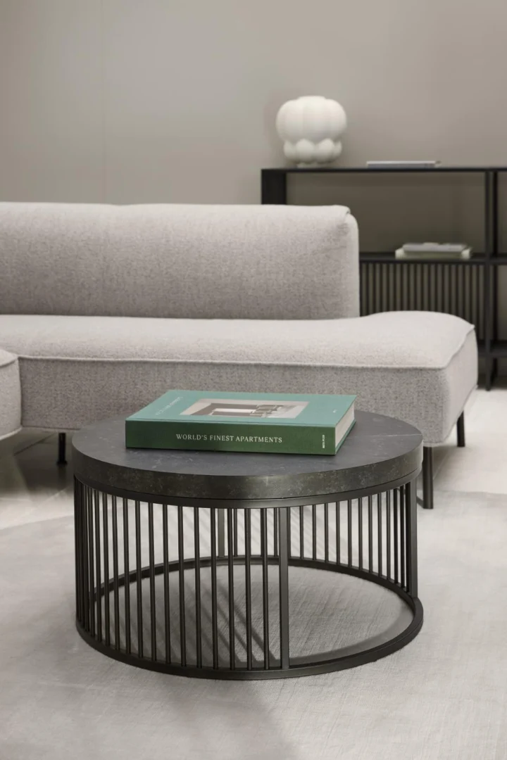 Een moderne woonkamer met de Salontafel Zwart 65 CM Rond Kunststof salontafel, een boek met de titel Worlds Finest Apartments erop, een lichtgrijze bank en een decoratieve plank met een witte vaas op de achtergrond.