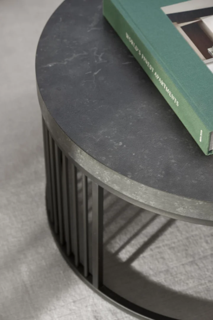 Een Salontafel Zwart 65 CM Rond Kunststof met verticale metalen lamellenpoten houdt het groene boek "Worlds Finest Apartments" vast op een lichtgrijze vloer met structuur.