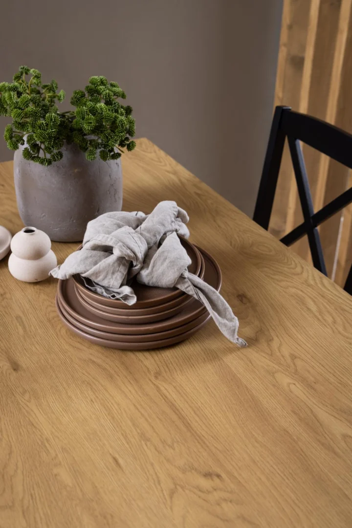 De Eettafel Nieuwe Sfeer 80 x 75 CM Rond, gemaakt van eikenhout en staal, is vormgegeven met gestapelde bruine borden, een geknoopt grijs kleed, een groene plant in een grijze vaas en twee kleine witte keramiekjes naast een zwarte stoel.