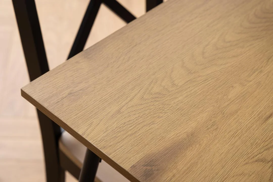 Close-up van de Eettafel Nieuwe Sfeer 80 x 75 CM Rond, gemaakt van eikenhout en staal, met de lichtbruine houtnerf. Een deel van een zwarte stoel met kruisrug is zichtbaar naast de tafel.